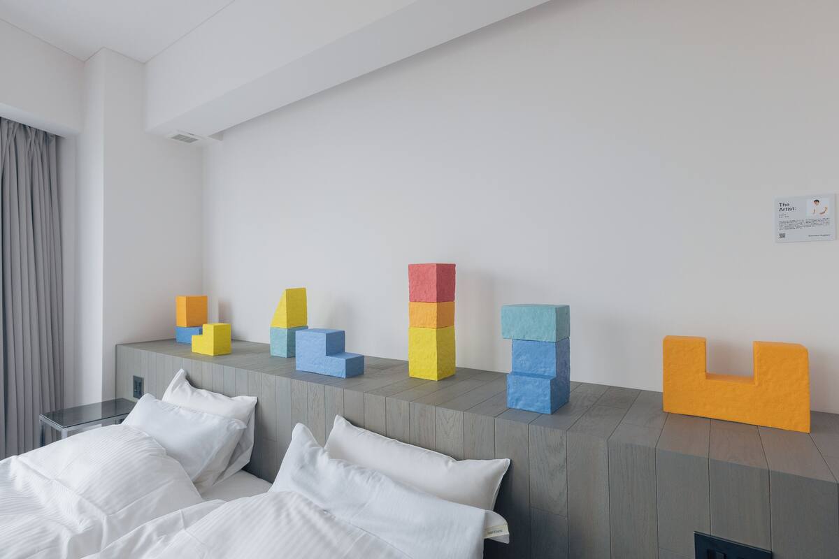 Ein Hotelzimmer mit einem Bett, einem Nachttisch, einem Wandbild und einem Stapel bunter Blöcke auf einer Konsole.