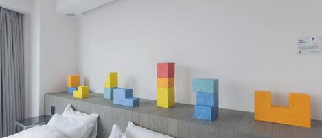 A hotel room with a bed, a bedside table, a wall-mounted artwork, and a stack of colorful blocks on a console table.