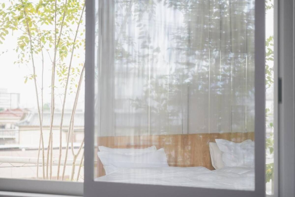 Ein Bett mit weißer Bettwäsche, einem Holz-Kopfteil und Blick auf Bäume durch transparente Vorhänge.