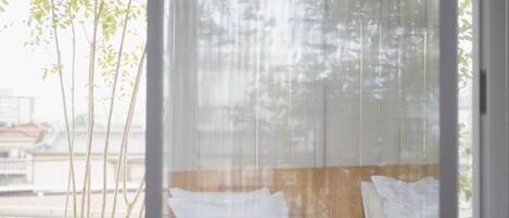 A bed with white bedding, a wooden headboard, and a view of trees through sheer curtains.