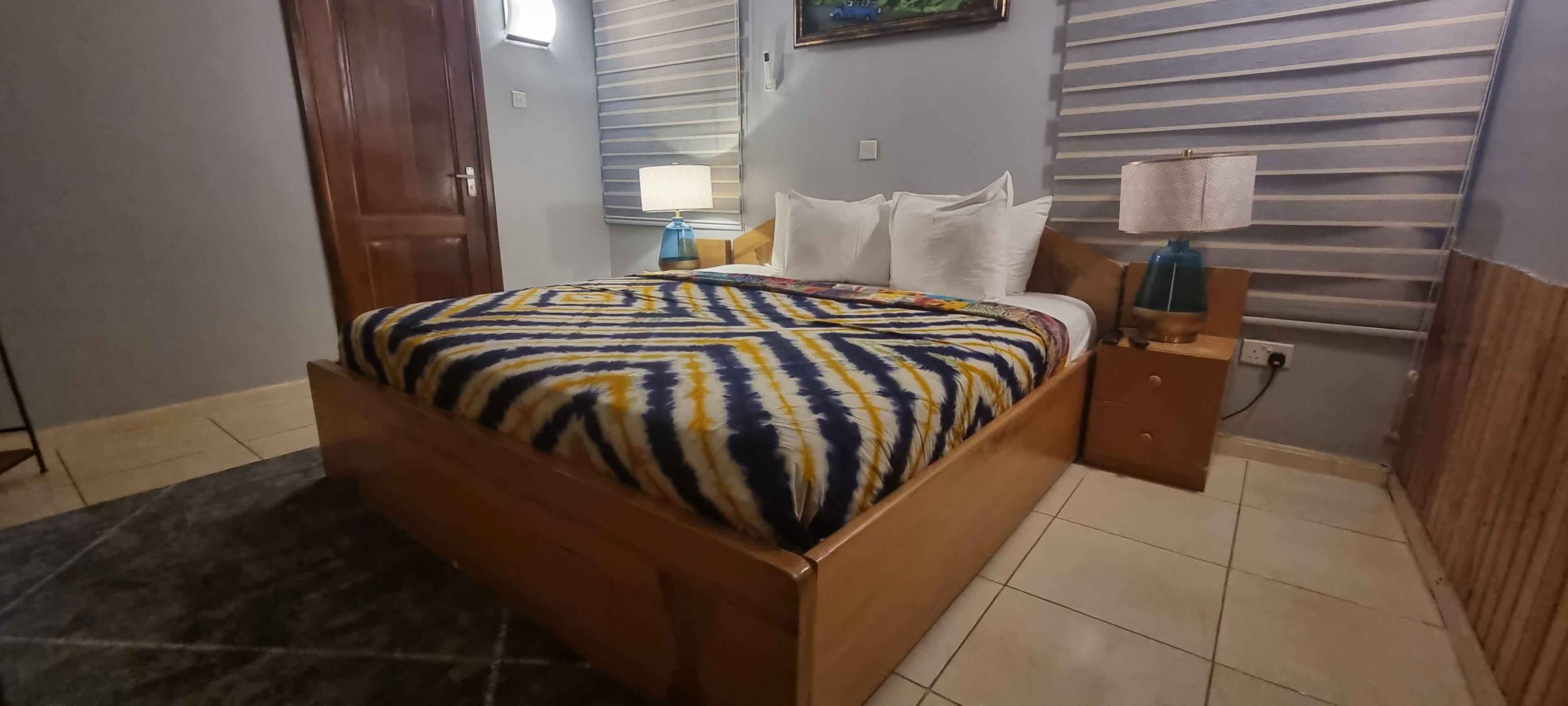 A bedroom with a bed, a nightstand, a lamp, a window with blinds, and a door.