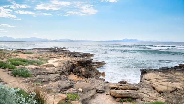 Uma costa rochosa com ondas, montanhas ao longe e céu claro.