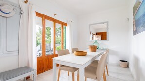 Uma sala de jantar com mesa branca, cadeiras bege, um banco de madeira e vista através de uma porta de vidro.