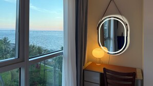 Habitación con escritorio, silla y espejo con luces. Una ventana ofrece vista al mar y follaje.