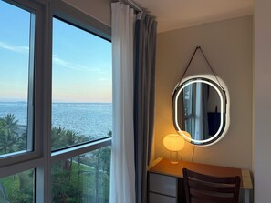A room with a desk, chair, and a mirror with lights. A window offers a view of the sea and greenery. - Playful 1BR Bayview Near Okada & MOA (Parañaque)