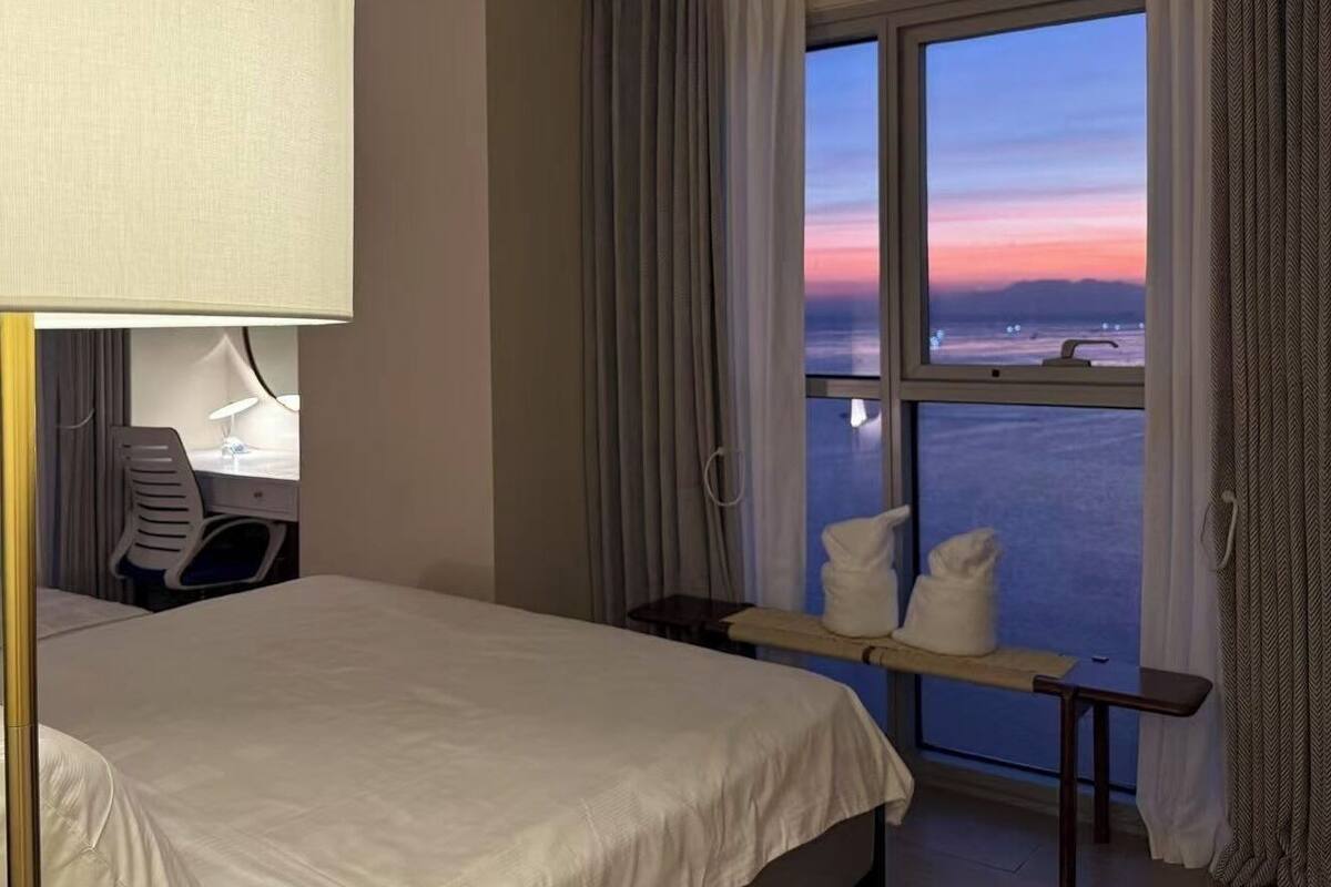 A hotel room with a bed, a desk, a chair, a window with curtains, and a view of the sea.