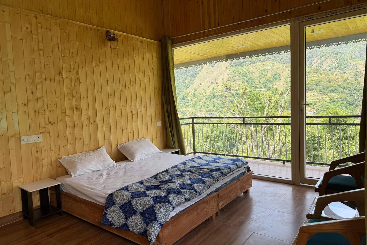 Quarto de madeira com cama, mesa de cabeceira e vista para uma paisagem montanhosa através de uma varanda.