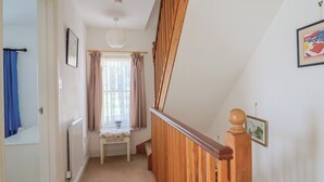 A narrow hallway with a wooden staircase, a radiator, a small table with a floral cushion, and a window with curtains. - Morpeth (Chard)