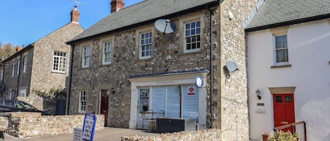 A stone building with a shop named 'The Village Shop' on the ground floor, featuring a signboard with various services.
