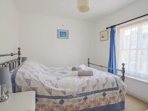 Un dormitorio con cama, mesita de noche, lámpara, ventana con cortinas y dos cuadros en la pared.