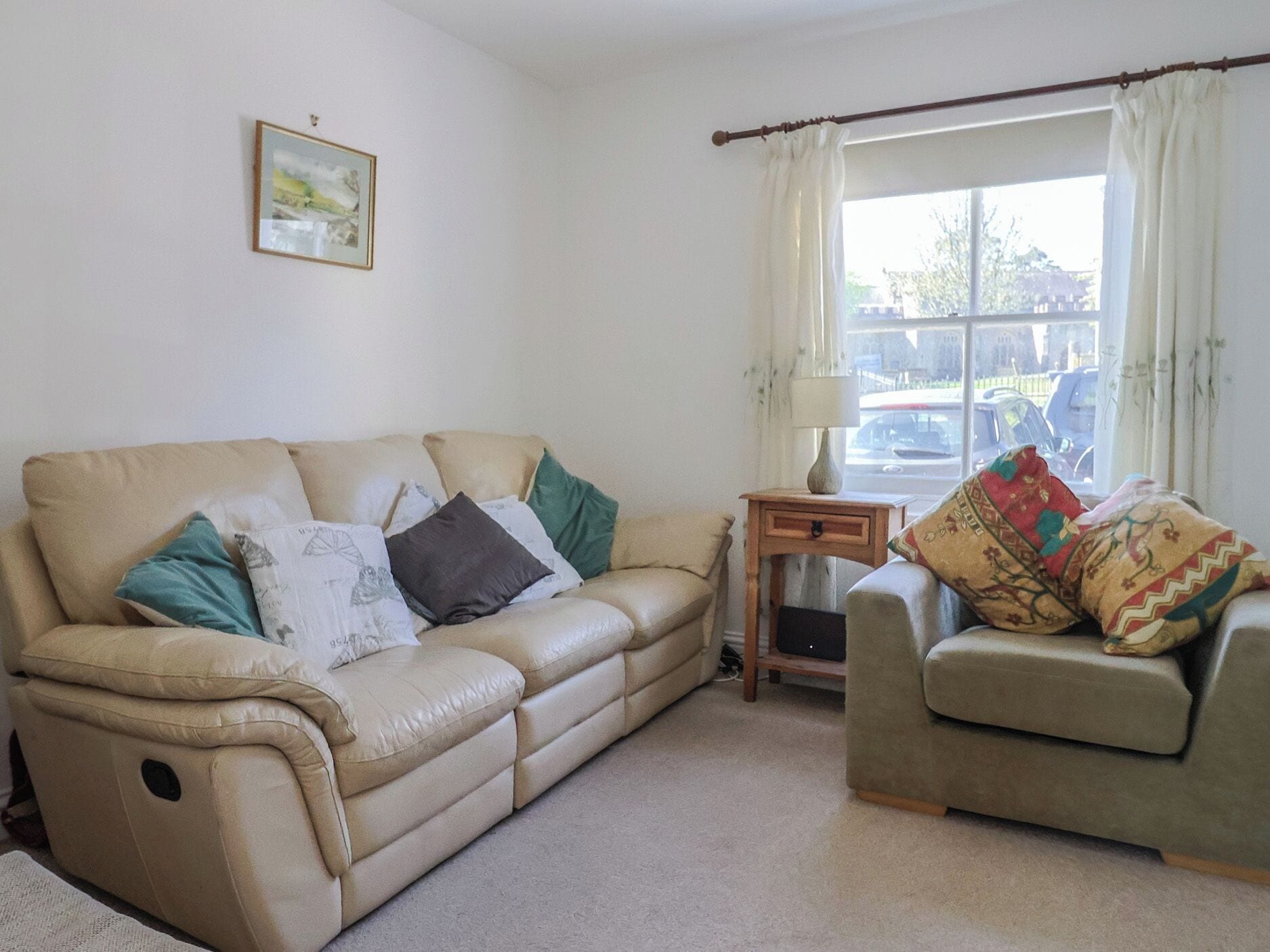 A living room with a beige sofa, a smaller armchair, a side table with a lamp, a framed picture on the wall, and a window with curtains.