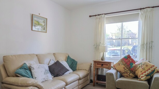 A living room with a beige sofa, a smaller armchair, a side table with a lamp, a framed picture on the wall, and a window with curtains. - Morpeth (Chard)