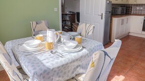 A dining table set with plates, cups, and cutlery, chairs with patterned cushions, a kitchen with cabinets and appliances, and a door leading to another room. - Morpeth (Chard)