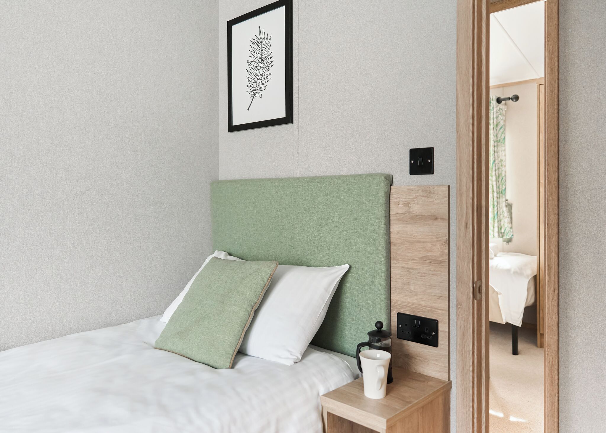 A neatly made bed with a green headboard, two pillows, a bedside table with a white mug, and a framed leaf artwork on the wall.