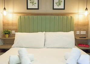 Ein Hotelzimmer mit einem Bett, zwei Kissen, einem grünen Kopfteil, Nachttischen mit Lampen, einer Zimmerpflanze, einer Fernbedienung und einem Buch auf einem der Tische.