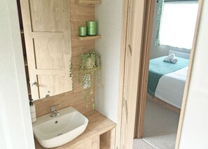 A bathroom with a wooden vanity, a sink, and a mirror. There is a green plant hanging on the wall and a window with a view of a bed. - Exclusive Lodge 4 bed HT (Landford, Nr Salisbury)