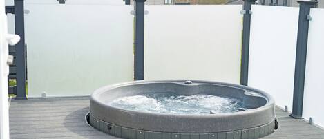 Un jacuzzi en una terraza de madera con una valla blanca y casas al fondo.
