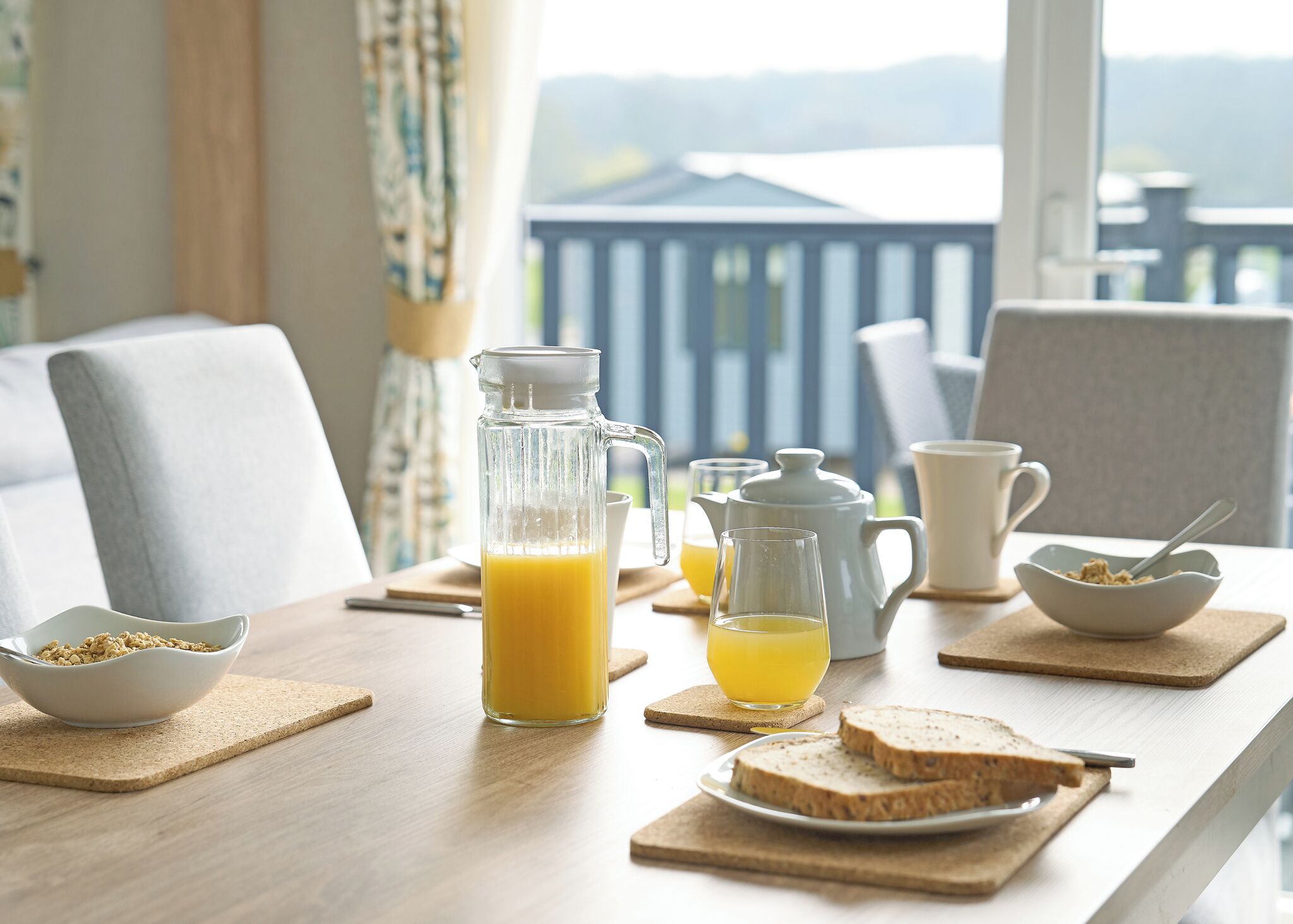 Une table mise pour le petit-déjeuner avec une carafe de jus d’orange, deux verres, une théière, un bol de céréales et des tranches de pain.