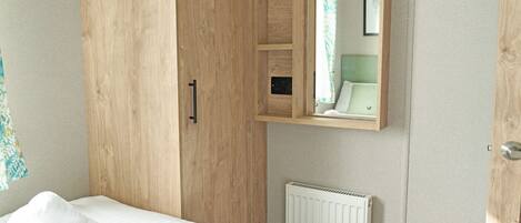 A bedroom with a wooden wardrobe, a bed with white bedding, a radiator, and a mirror reflecting a bathroom.