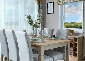 A dining table with chairs, a vase with greenery, wine glasses, and a view of a landscape through a window. - Exclusive Lodge 3 bed HT (Hayle)