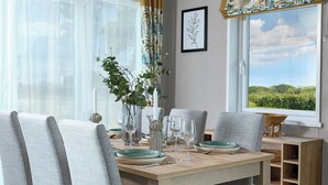 Une table à manger avec des chaises, un vase orné de verdure, des verres à vin et une vue sur un paysage par la fenêtre.