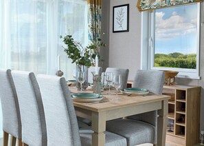 A dining table with chairs, a vase with greenery, wine glasses, and a view of a landscape through a window. - Exclusive Lodge 3 bedroom Hot Tub sleeps 6 (Hayle)
