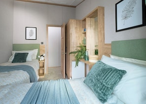 A bedroom with two beds, a wooden wardrobe, a radiator, and a framed leaf artwork. - Exclusive Lodge 3 bed HT (Hayle)