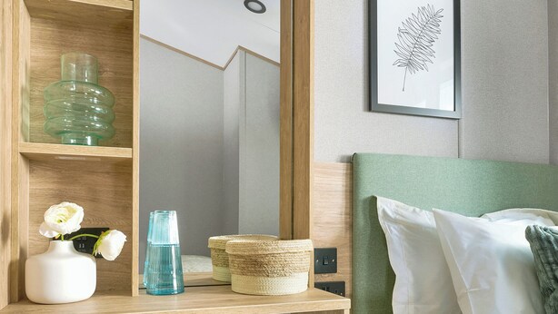 Une chambre d’hôtel avec une étagère en bois, un lit doté d’une tête de lit verte, des oreillers blancs, une œuvre d’art représentant une feuille encadrée et un vase blanc contenant des fleurs.