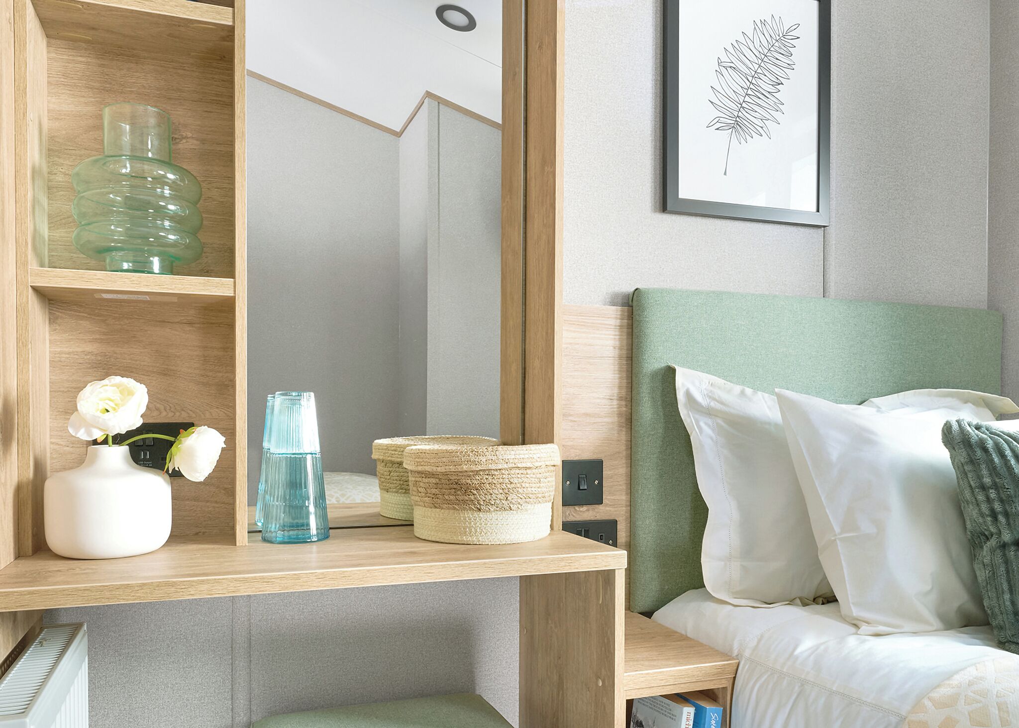 A bedroom with a green headboard, a bedside table with a vase of white flowers, a blue glass bottle, and woven baskets.