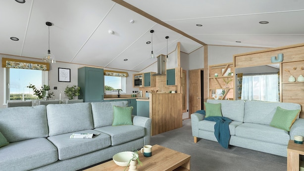 A modern living room with two grey sofas, a wooden coffee table, and a kitchen area with wooden cabinets and a window. - Exclusive Lodge 3 bed HT (Great Yarmouth)