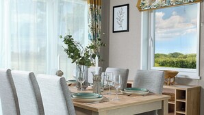 A dining area with a wooden table, grey upholstered chairs, and a view of greenery through the window. - Exclusive Lodge 3 bed HT (Great Yarmouth)