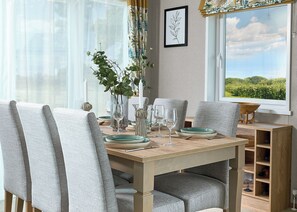 A dining area with a wooden table, grey upholstered chairs, and a view of greenery through the window. - Exclusive Lodge 3 bed HT (Great Yarmouth)