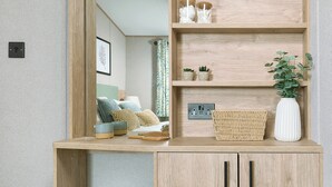 A room with a wooden desk, a mirror, a bed with pillows, a small shelf with decorative items, and a green bench. - Exclusive Lodge 3 bed HT (Great Yarmouth)