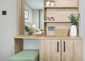 A room with a wooden desk, a mirror, a bed with pillows, a small shelf with decorative items, and a green bench. - Exclusive Lodge 3 bed HT (Great Yarmouth)