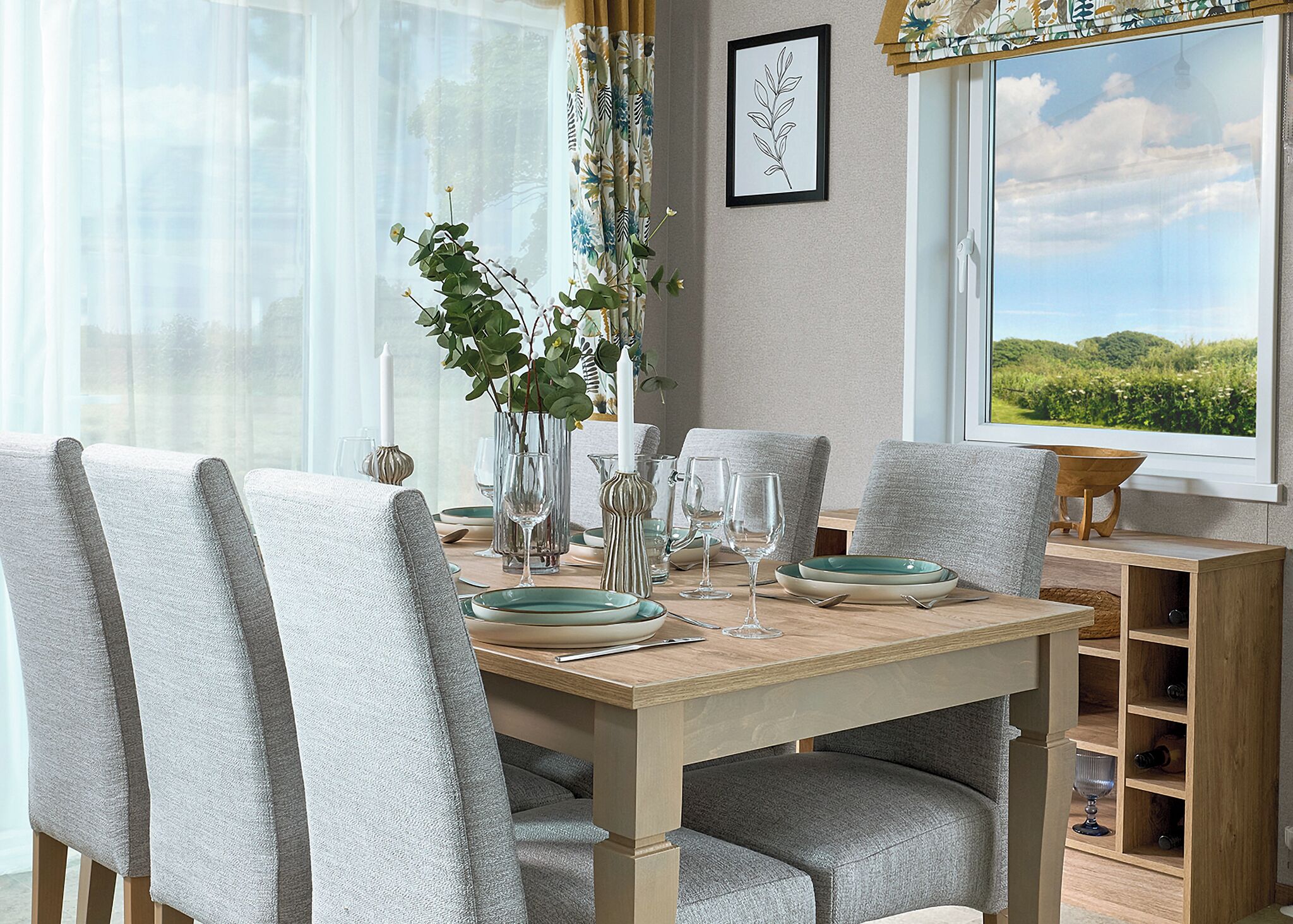 A dining table with chairs, a vase with greenery, wine glasses, and a framed picture on the wall.