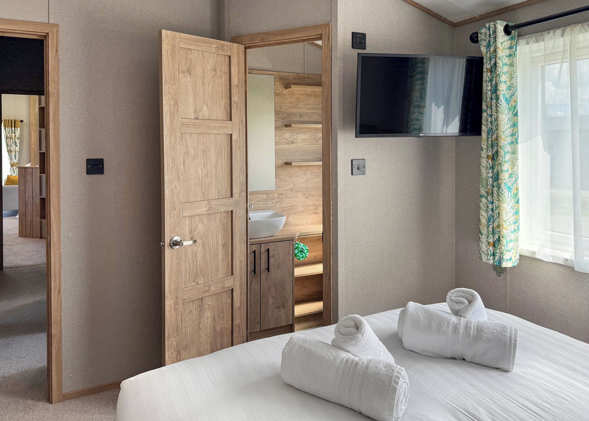 A bedroom with a bed, two towels, a TV, a wooden wardrobe, a mirror, and a window with curtains.