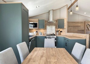 A modern kitchen with teal cabinetry, a wooden dining table, and pendant lighting. - Exclusive Lodge 4 bed HT Pet (Great Yarmouth)