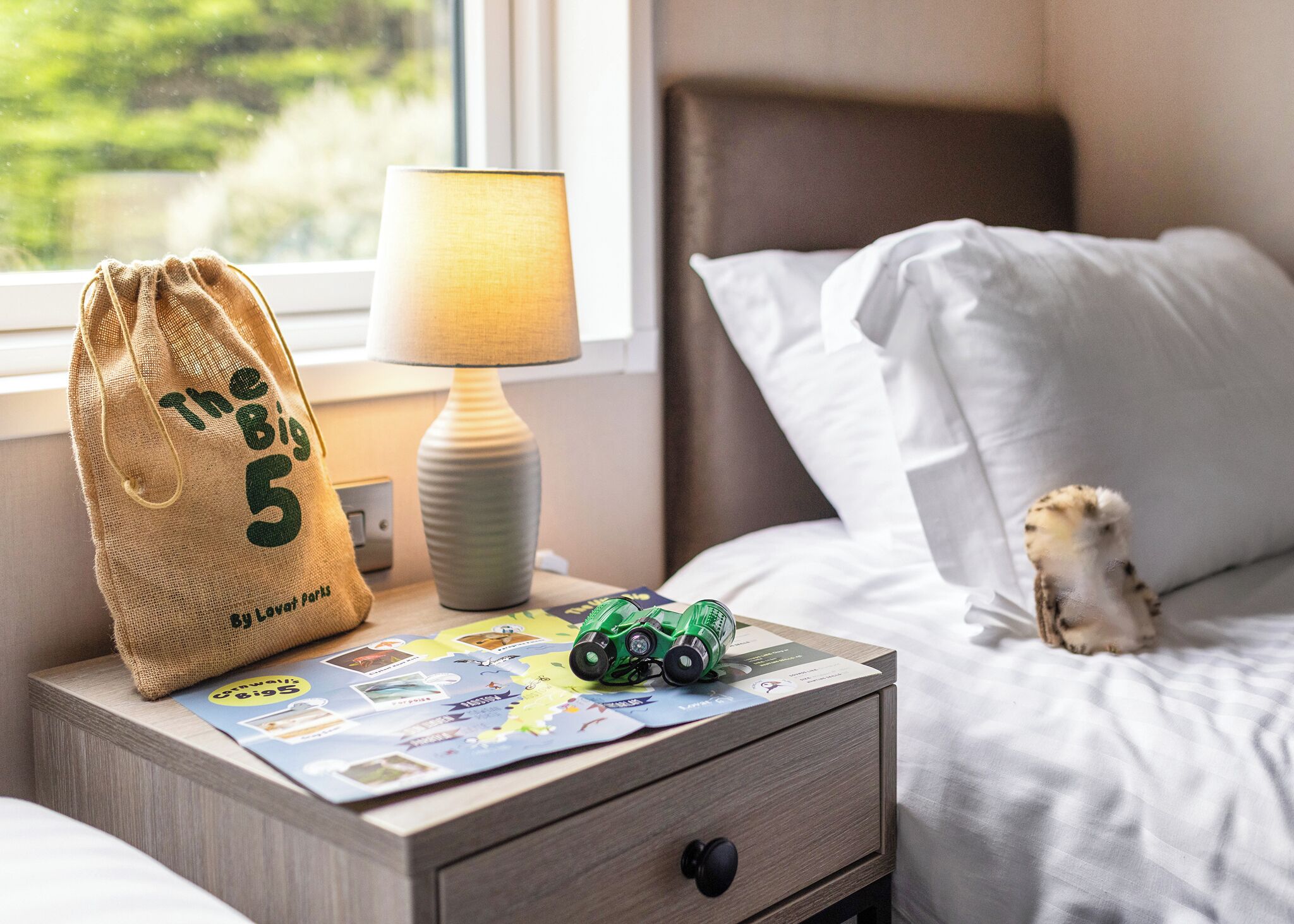 Een netjes opgemaakt bed met witte lakens, een lamp, een kleine tas met tekst, een autootje en een kaart op een nachtkastje.