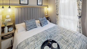 Una cama bien tendida con un edredón a rayas, dos almohadas, mesitas de noche con lámparas, una bandeja con dos tazas y cuadros en la pared.