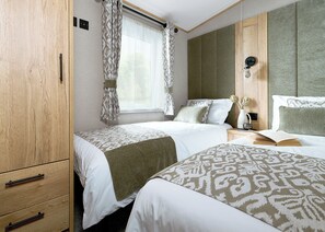 A hotel room with two beds, a wooden wardrobe, a window with patterned curtains, and a bedside table with a book and a vase. - 3 bedroom accommodation in Great Yarmouth (Great Yarmouth)