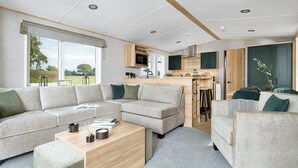A modern living room with a sofa, ottoman, and coffee table, a kitchen area with bar stools, and a view of the outdoors. - Premium Holiday Home 3 bed HT Pet (Great Yarmouth)