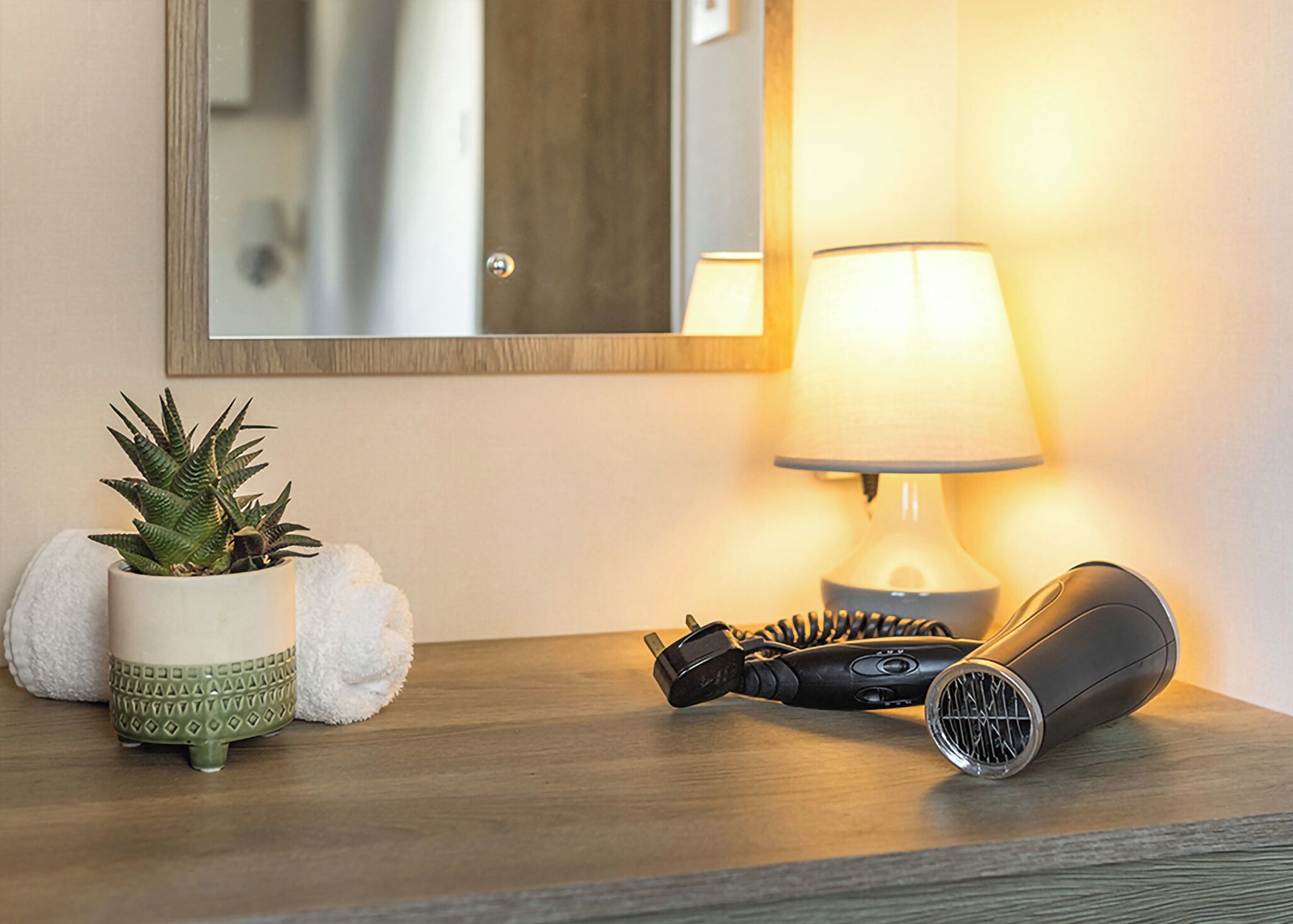 A wooden bedside table with a lamp, a coiled hair dryer, and a potted plant.