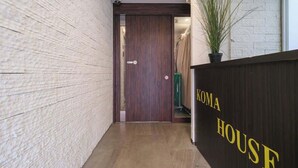 Um corredor com uma porta de madeira, uma mesa de recepção com a placa 'KOMA HOUSE' e um vaso de planta.