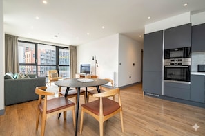 A modern kitchen with built-in appliances, a dining area with a round table and wooden chairs, and a sofa with a city view. - Modern Meets Urban Living (London)