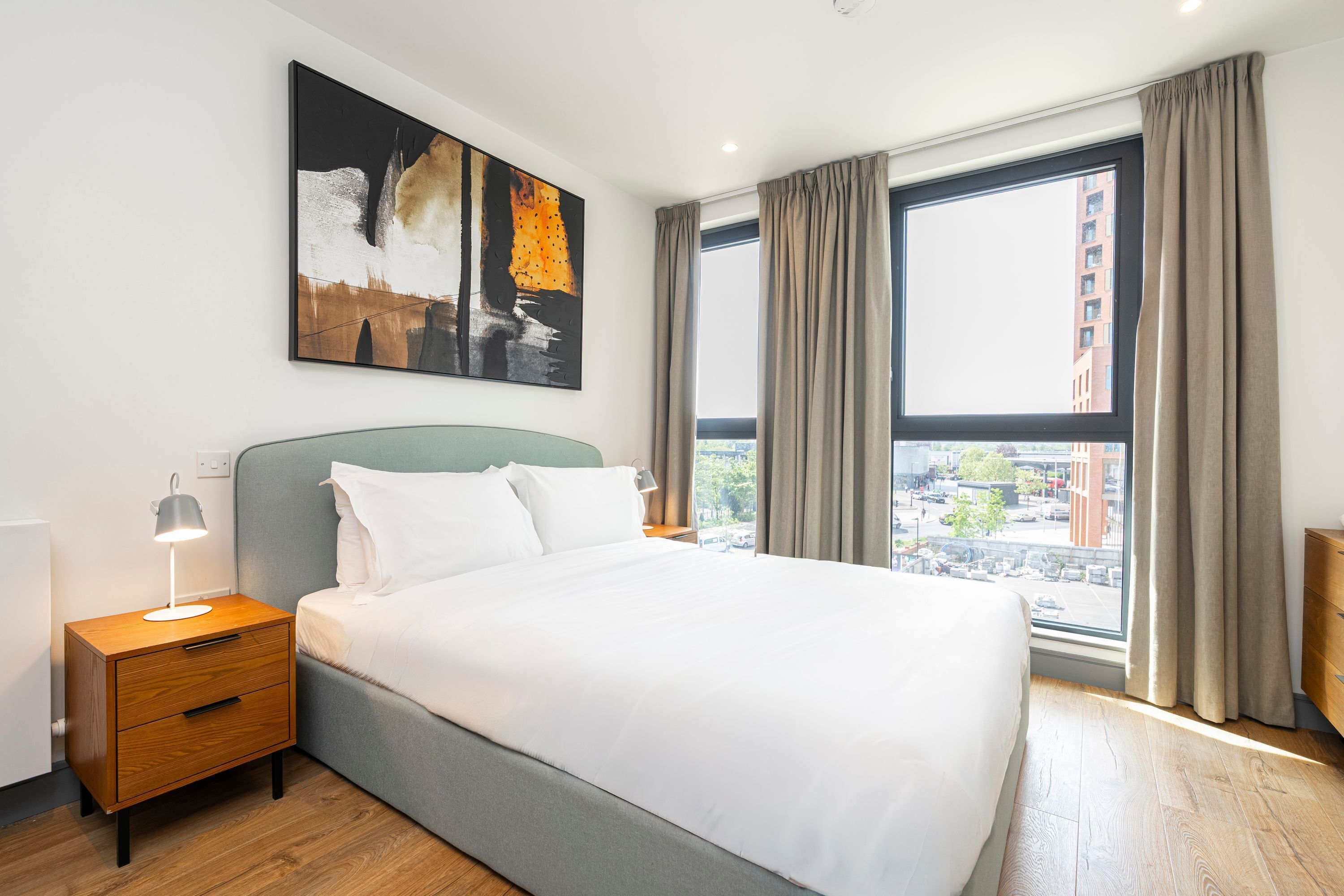 A hotel room with a bed, a nightstand, a lamp, a painting, and a window with curtains.