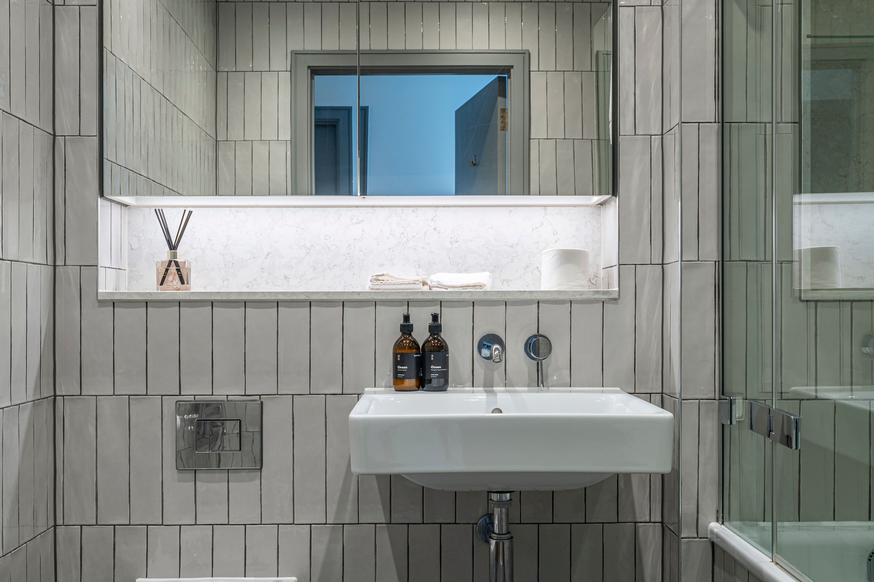 A bathroom with a white sink, a mirror above it, and a window. There are two bottles on the sink.