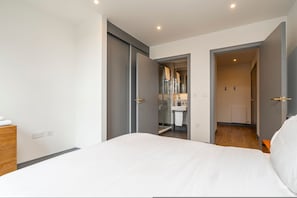Ein Schlafzimmer mit Bett, einem Holzkonsolenmöbel, einem Badezimmer mit Duschkabine und einem Flur, der zu einem weiteren Raum führt.