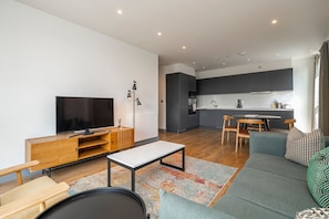 Ein modernes Wohnzimmer mit Fernseher, Sofa und Couchtisch, eine Küche mit dunklen Schränken und ein Essbereich mit Stühlen und einem Tisch.