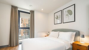 Un letto rifatto con cura in una camera d'albergo moderna, con un'ampia finestra, un comodino con lampada e due stampe botaniche incorniciate al muro.