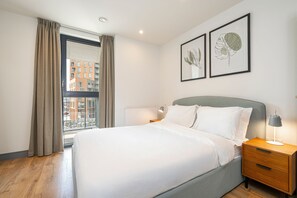 Ein ordentlich bezogenes Bett in einem modernen Hotelzimmer mit großem Fenster, einem Nachttisch mit Lampe und zwei gerahmten Pflanzenbildern an der Wand.